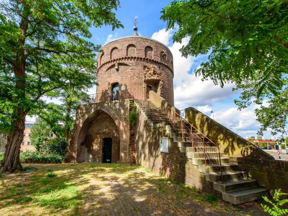 De middeleeuwse Rattentoren in Roermond, omgeven door groene bomen, met een brede trap naar de ingang op de eerste verdieping en een boogvormige doorgang op de begane grond.