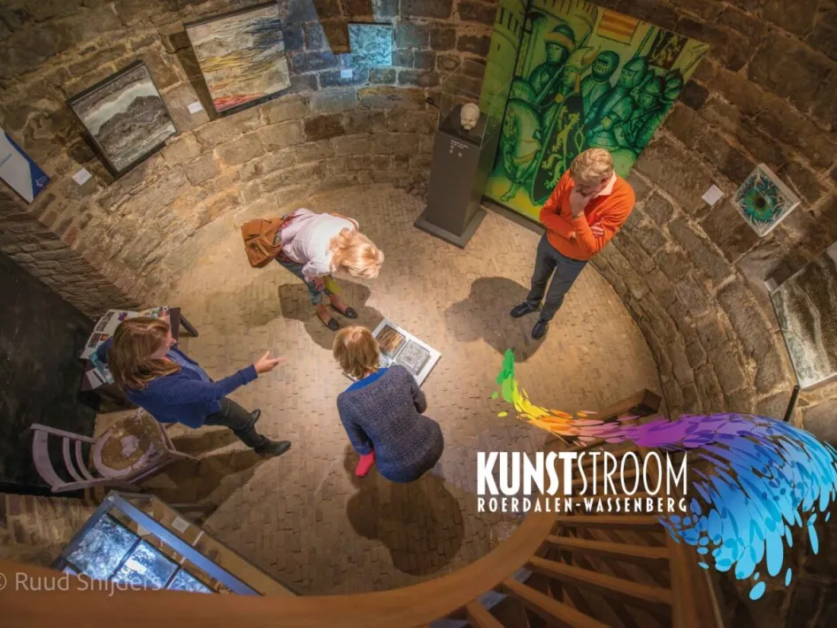 Bezoekers bekijken kunstwerken in een ronde stenen toren tijdens Kunststroom Roerdalen-Wassenberg. Op de vloer liggen kunstwerken en schilderijen hangen aan de muren.