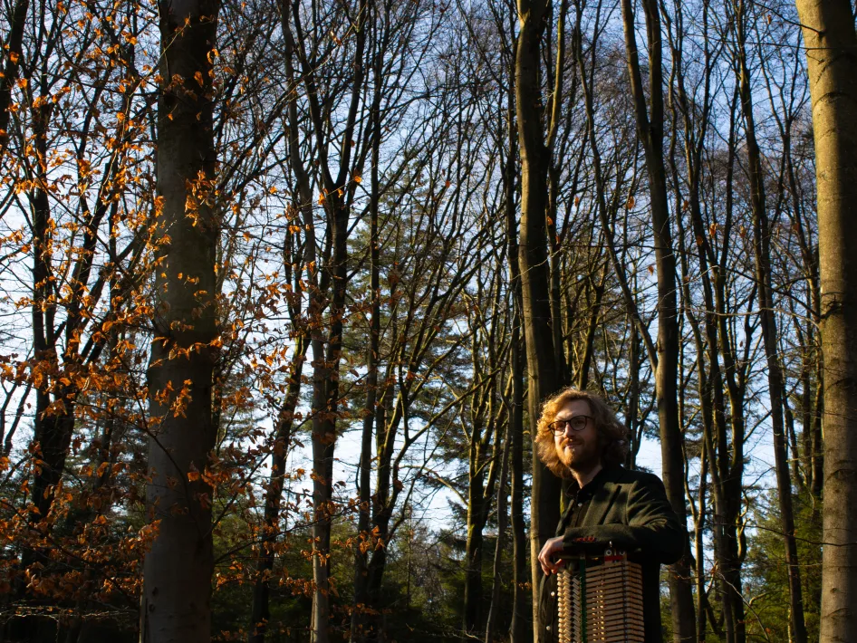 Persoon met lange haren staat tussen hoge bomen in een bos, met zonlicht dat door de takken valt en herfstbladeren aan de bomen.