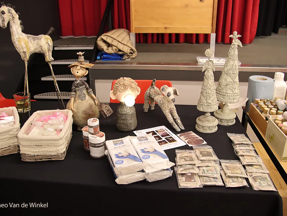 Tafel met creatieve handgemaakte decoraties, waaronder beelden van een paard, poppen, kerstbomen en een hond, omringd door verfmaterialen, klei en informatiefolders tijdens een hobbymarkt.