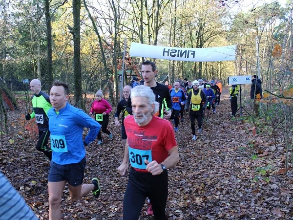 Groep hardlopers rent door een bosrijk parcours tijdens een natuurloop, onder een wit 'Finish'-spandoek in herfstachtige omgeving.
