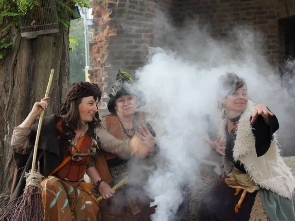 Drie vrouwen in theatrale heksenkostuums poseren tussen rookwolken tijdens een buitenvoorstelling, passend bij de sfeer van de lezing Heksenwaan.