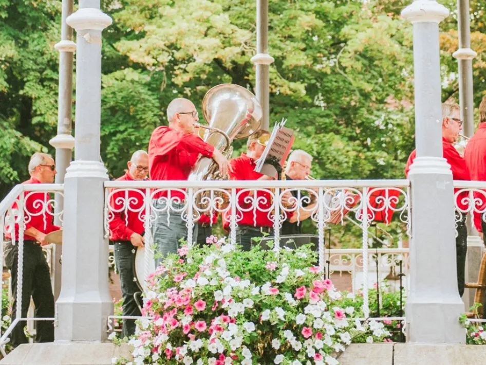 Kioskconcerten Munsterplein met muzikanten in rode kleding die optreden in een muziekkiosk, omringd door bloemen en groen.