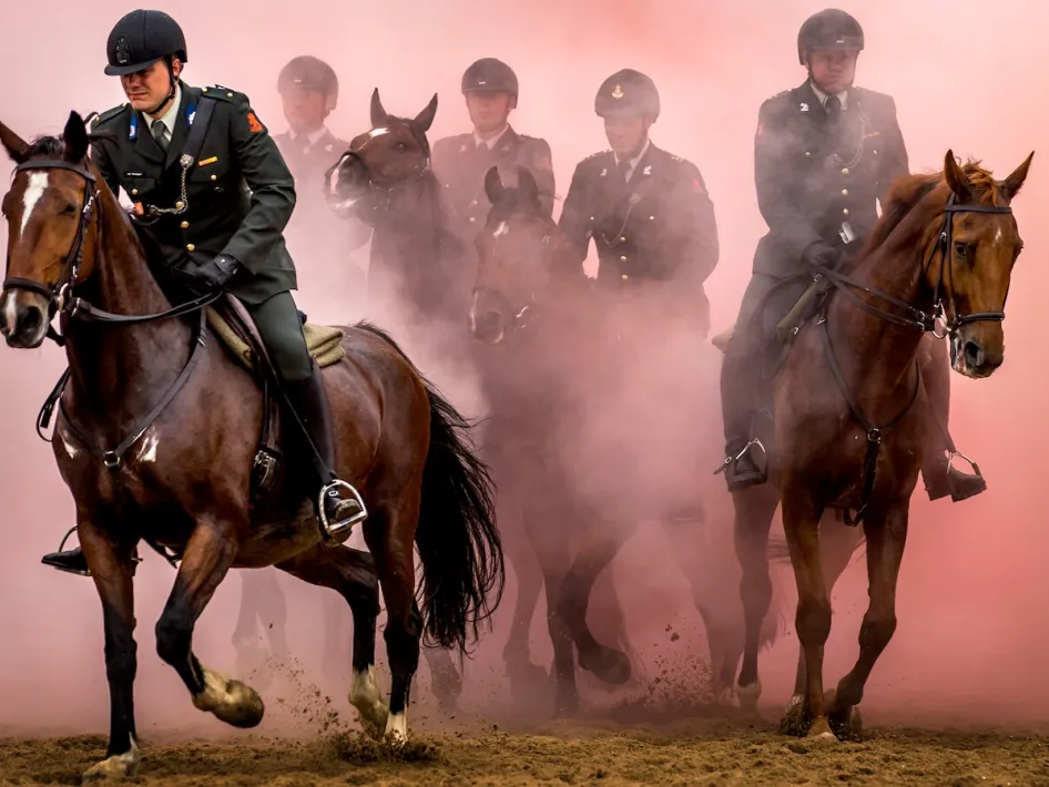 Groep militairen te paard rijdt door een wolk van rood rook tijdens een demonstratie, gehuld in uniform en helmen.