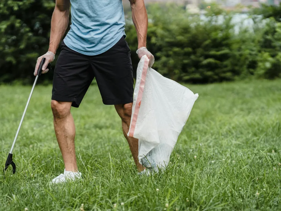 Vrijwilliger raapt zwerfafval op in het gras met een grijper en een afvalzak tijdens een opruimactie in de natuur.