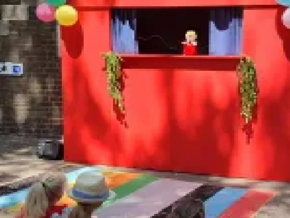Kinderen kijken naar een poppenkastvoorstelling bij een rood theaterdecor buiten tijdens het Vertel- en poppentheater.
