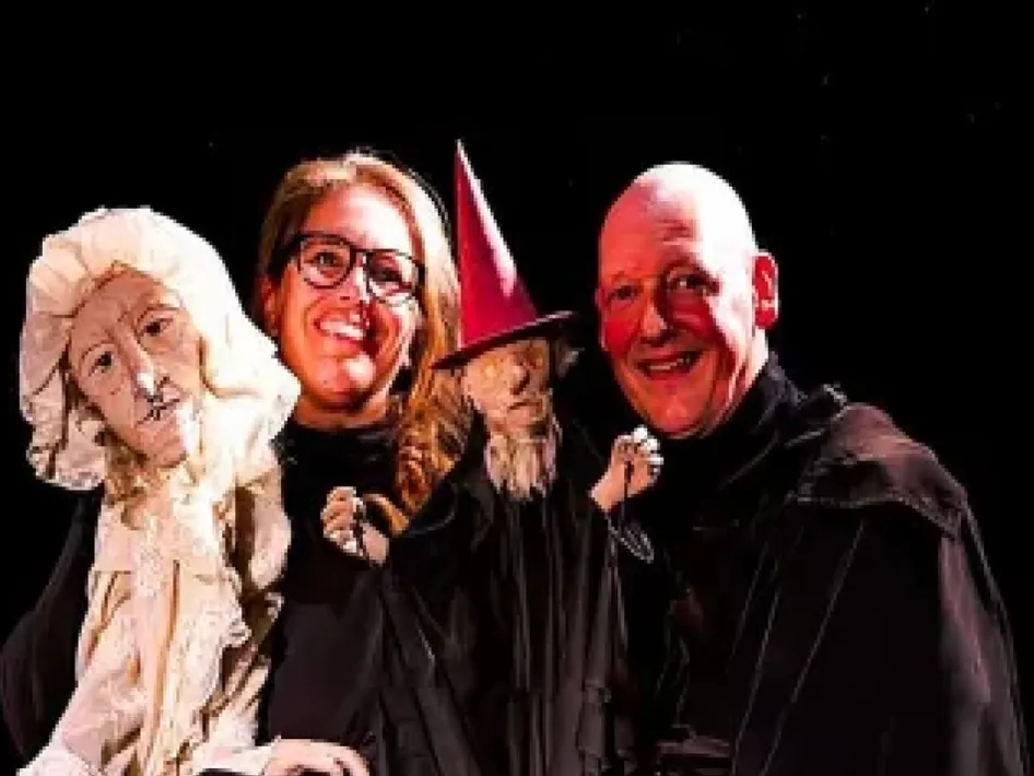 Twee poppenspelers met handpoppen, waaronder een heks met rode hoed, tegen een donkere achtergrond, tijdens het Beleef het vertel- en poppentheaterfestival bij de Rattentoren in Roermond. Geniet op 19 april van gratis voorstellingen vol verhalen en poppenspel .Vertel- en poppentheaterfestival
