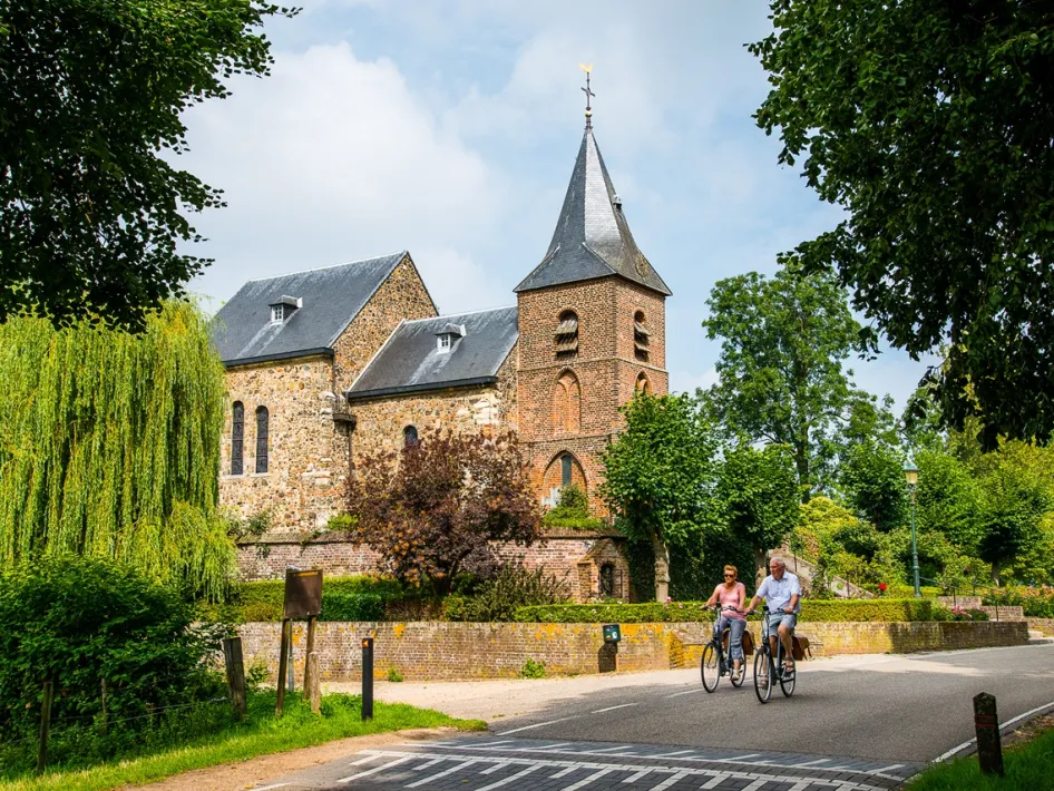 Fietsers rijden langs een landelijke weg bij een kerk in Limburg, omringd door groen en bomen op een zonnige dag.