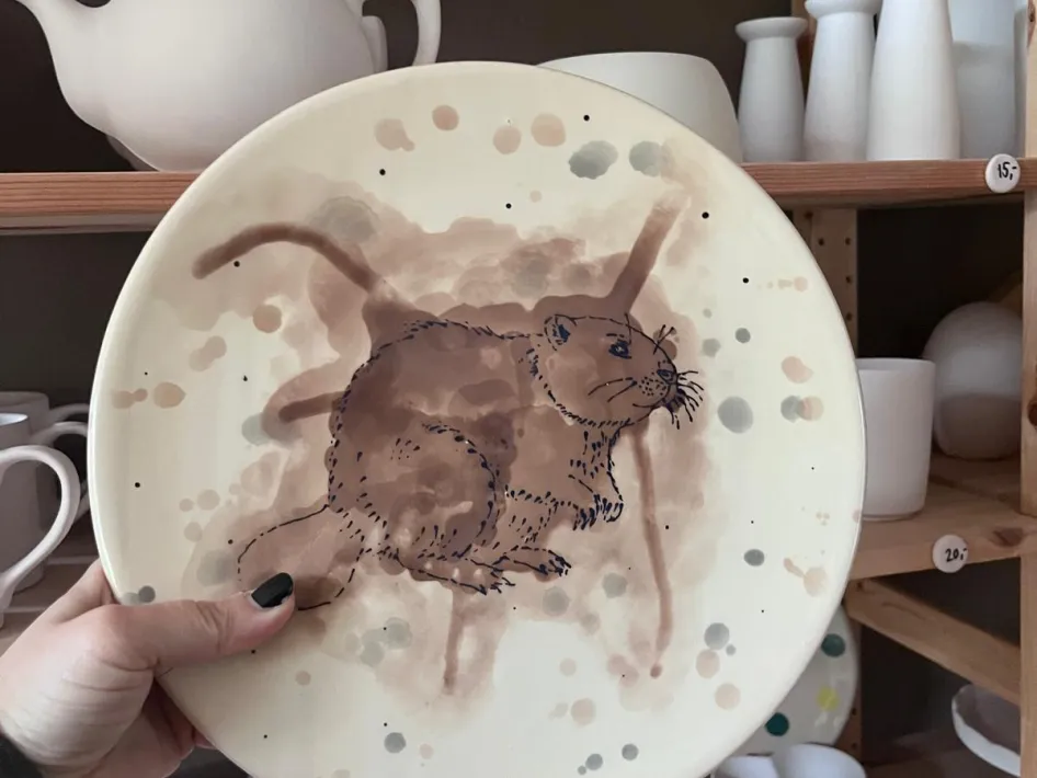 Hand houdt een beschilderd Keramiek bord met een bruine beverillustratie vast, voor een plank met onbeschilderde kopjes, vazen en theepotten in een atelier.