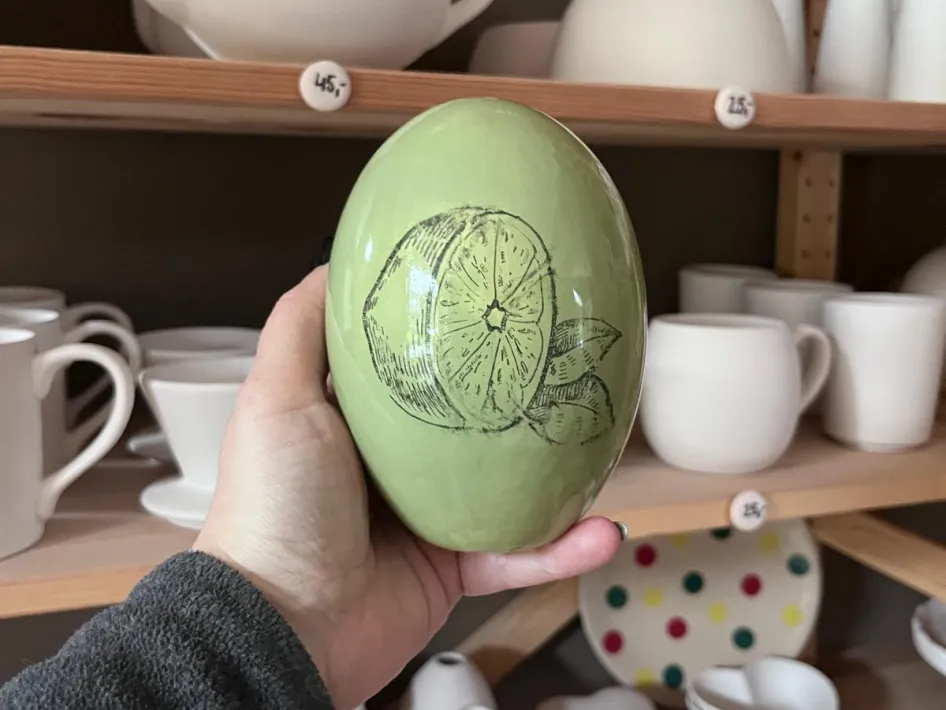 Hand houdt een groen beschilderd Keramiek vaasje met een citroenillustratie vast, voor een plank met onbeschilderde kopjes, theepotten en schaaltjes in een atelier.