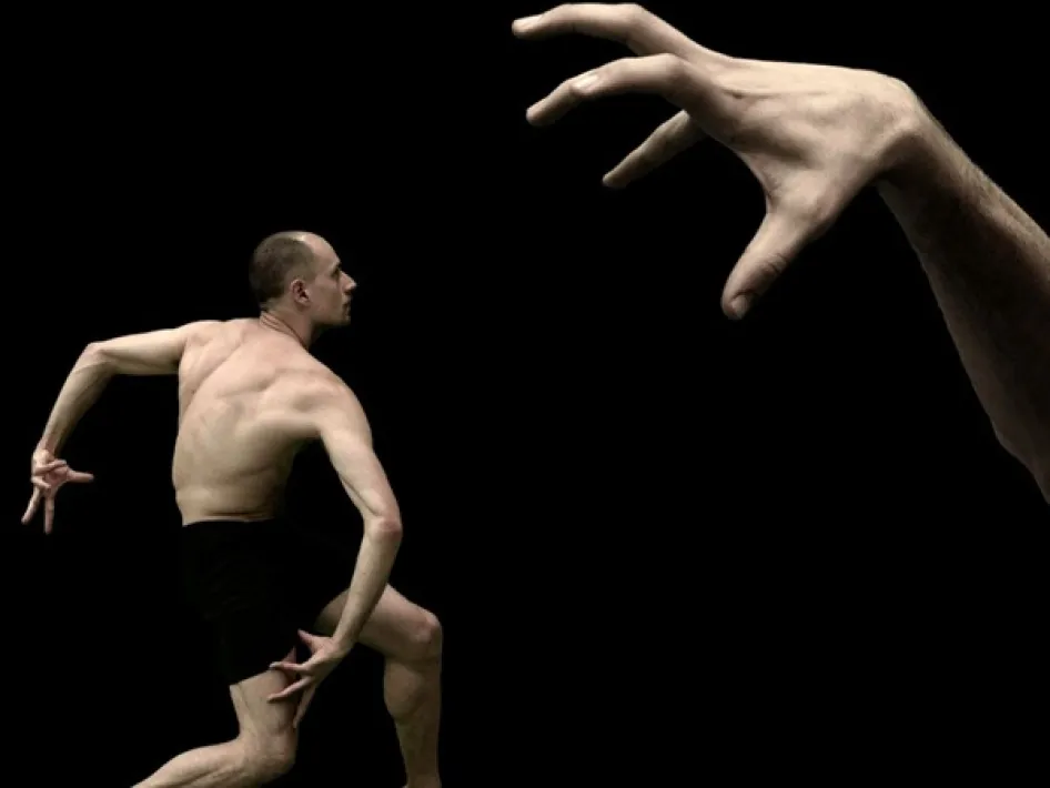 Danser in beweging tijdens theatervoorstelling silent gestures tegen een zwarte achtergrond terwijl een grote hand boven hem lijkt te grijpen