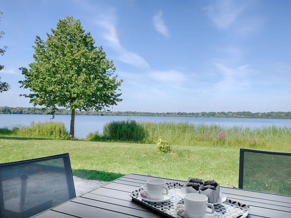 Een terras met een tafel en stoelen, uitkijkend over een groen veld en een groot meer op een zonnige dag