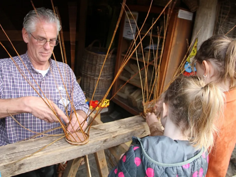 Kinderen leren takken vlechten tijdens een vlechtmiddag, terwijl een begeleider laat zien hoe je met wilgentakken een mand maakt.