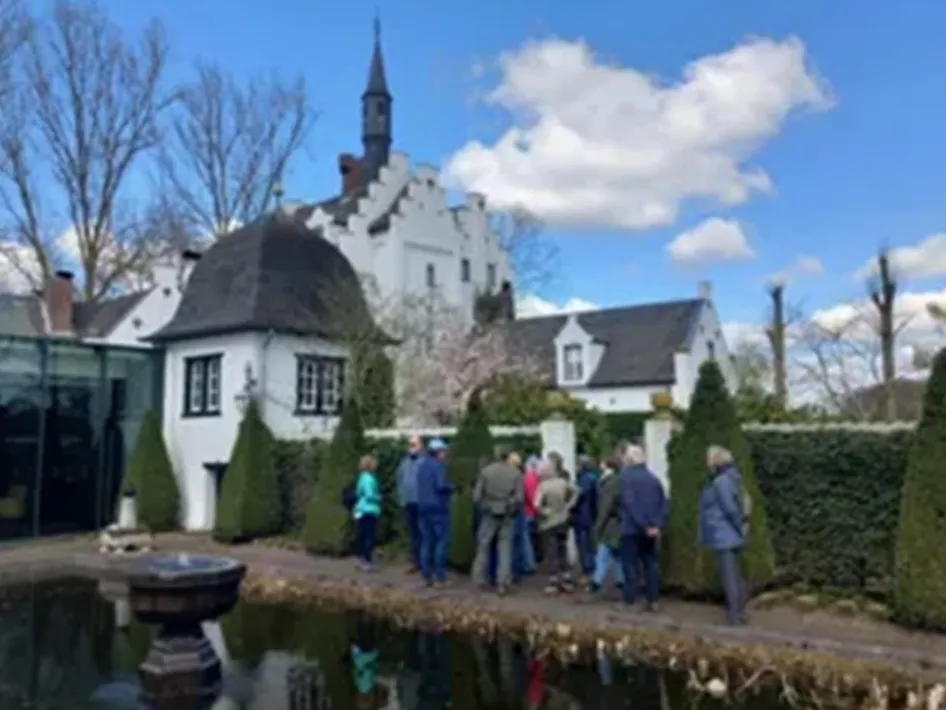 Groep mensen tijdens een wandeling langs een vijver bij een wit historisch gebouw met trapgevel en toren, omgeven door tuin en bomen onder een blauwe lucht