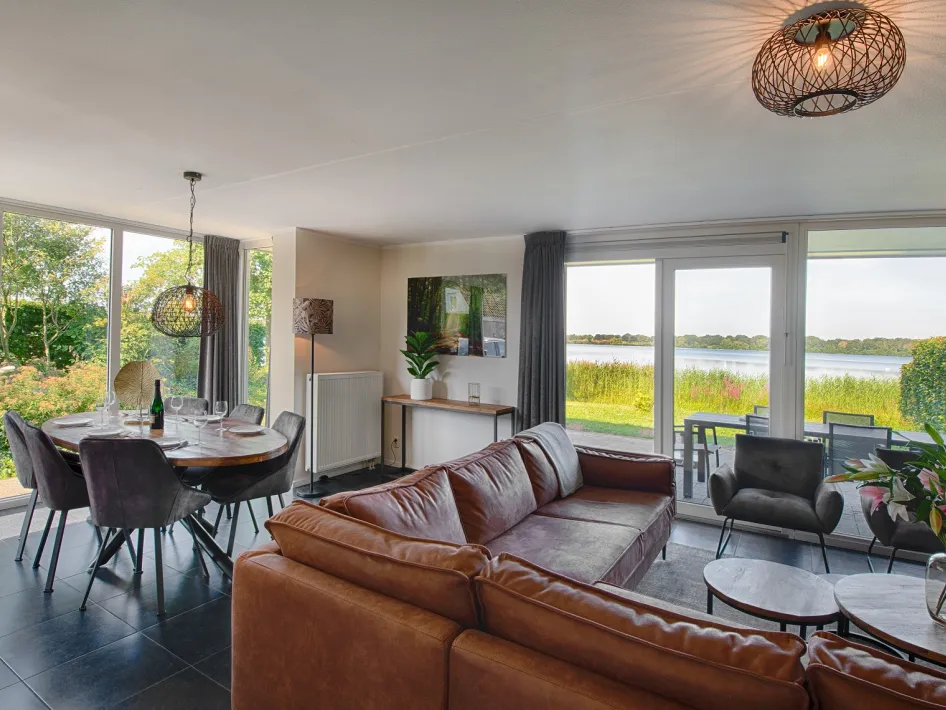 A cozy living room with a dining table and seating area, large windows with a view of a lake, and a terrace with garden furniture.