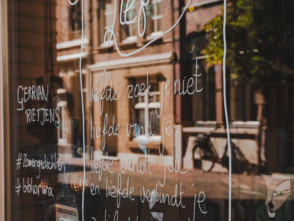 Etalageraam met een handgeschreven zomergedicht getiteld ‘Lief’ van Gerrian Rietjens, met hashtags #zomergedichten en #bibliorura, en weerspiegeling van de straat en gebouwen in Roermond.