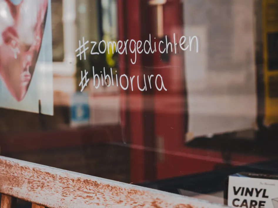 Etalageraam met handgeschreven hashtags #zomergedichten en #bibliorura, met op de achtergrond vage reflecties van posters en straatbeeld.