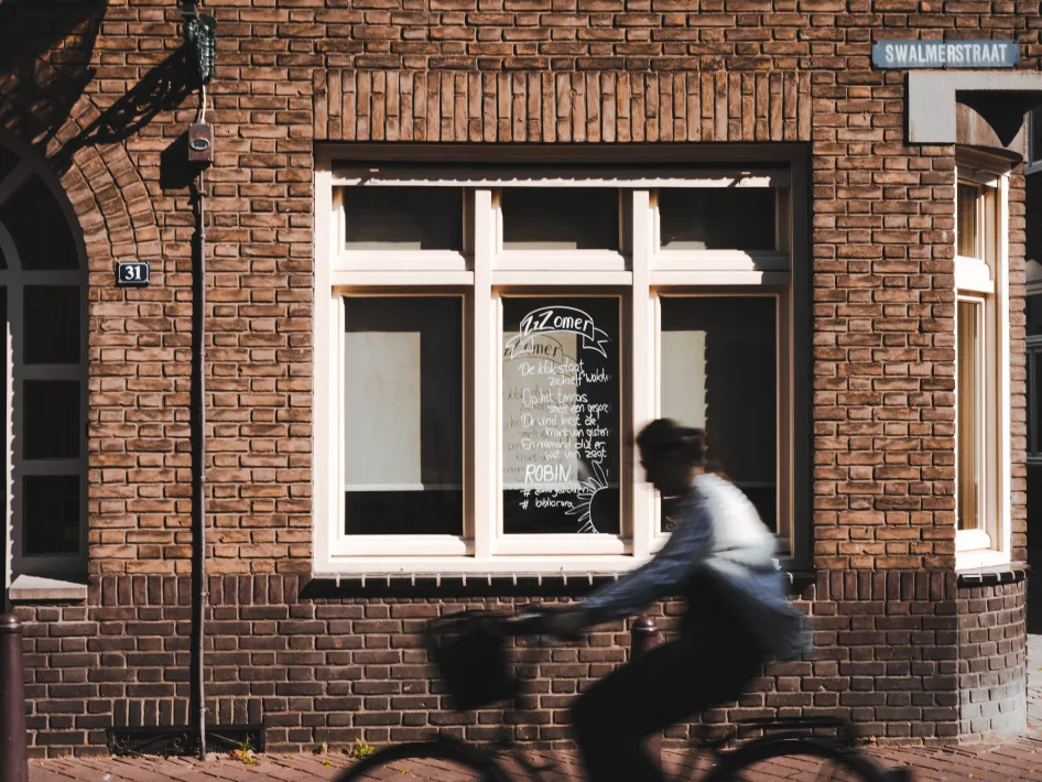 “Straatbeeld met een fietsende persoon voor een bakstenen gevel aan de Swalmerstraat in Roermond, met een zomergedicht op het raam van huisnummer 31.