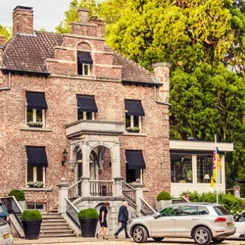 Vooraanzicht van Kasteeltje Hattem in Roermond, een klein kasteel met een klassieke uitstraling, omringd door groen en bijgebouwen, met geparkeerde auto’s en gasten op de trappen.