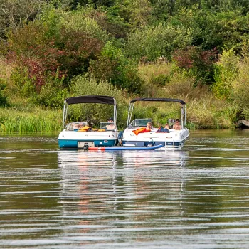 Twee motorboten en een opblaasbootje met mensen op een rustige rivier, omringd door groen en bomen.