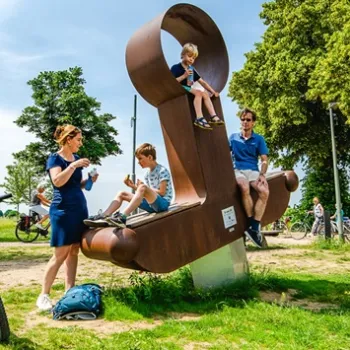 Fietsers en een gezin nemen pauze op en rondom een groot kunstwerk in de vorm van een zitobject langs de Maasfietsroute, omringd door bomen, fietsen en een zomers landschap.