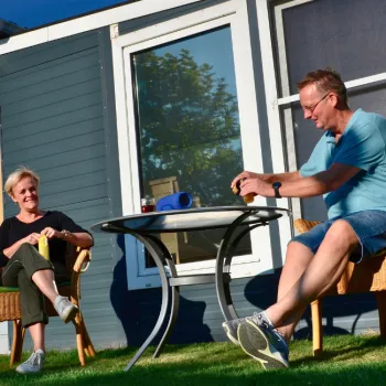 Man en vrouw ontspannen op een terras bij een chalet op een camping, omringd door groen en zonlicht.