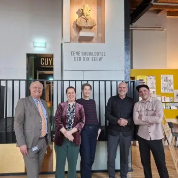 Groepsfoto van vijf lachende personen die de start van de voorbereiding van de terugkeer van de Koninklijke Troon vieren in de hal van het Cuypershuis in Roermond, onder een buste van architect Pierre Cuypers.