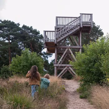 Twee kinderen wandelen over een zandpad richting een houten uitkijktoren, omringd door bomen en struiken.