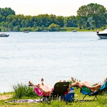Mensen relaxen op een grasstrand aan het water, met boten en zeilboot op de achtergrond op een zonnige dag in Limburg.