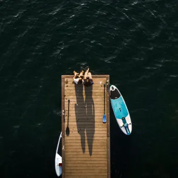 Luchtfoto van twee mensen die aan het einde van een houten steiger zitten boven donker water, met aan weerszijden een SUP-board en peddels in het water en op de steiger.