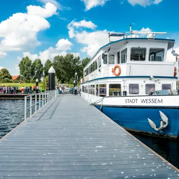 Rondvaartboot Stadt Wessem aangemeerd aan de steiger op de Maasplassen, waar bezoekers aan boord gaan voor de Asperge Cruise Voilà onder een blauwe lucht met wolken
