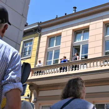 Mensen kijken vanaf een balkon naar een evenement op straat, terwijl voorbijgangers omhoog kijken naar het gebouw op een zonnige dag.