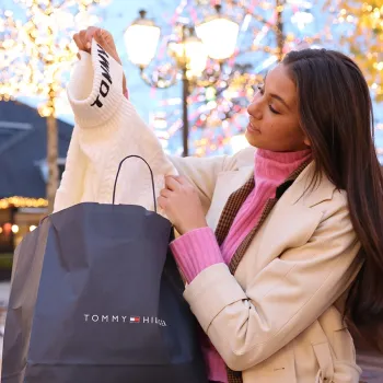 Frau holt einen weißen Pullover aus einer Tommy Hilfiger-Tasche während der Black Friday Days im Designer Outlet Roermond, umgeben von stimmungsvoller Weihnachtsbeleuchtung.