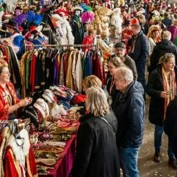 Een sfeervolle, drukke carnavalsmarkt in een grote hal met kraampjes vol kleurrijke carnavalskleding, accessoires en enthousiaste bezoekers. (AI gegenereerd)