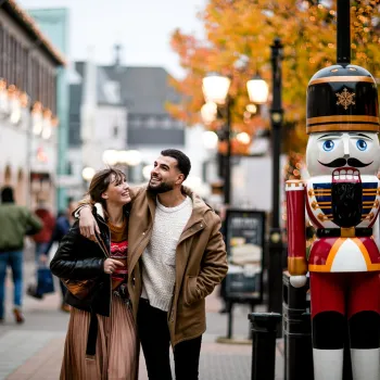 Stel wandelt lachend langs een notenkrakerfiguur tijdens de Christmas Sale bij Designer Outlet Roermond, omringd door feestelijke verlichting en herfstkleuren.