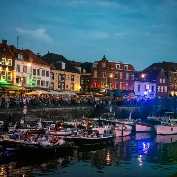 Drukke kade in Roermond tijdens het Roerdelta Concert, met toeschouwers op de kade en tientallen boten vol bezoekers op de Roer, tegen de achtergrond van sfeervol verlichte historische panden bij schemering.