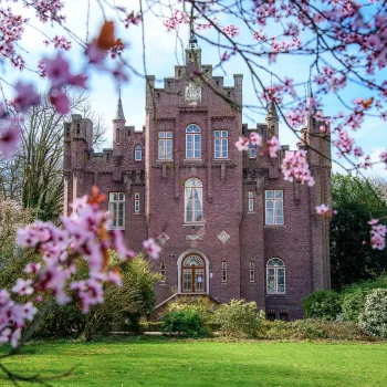 Kasteel Aerwinkel in Roerdalen, gezien door bloeiende roze takken in de voorgrond, met groene tuin en voorjaarslucht op de achtergrond.