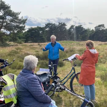 Stadsgids geeft uitleg aan deelnemers van de Fietstocht Rondje Weert tijdens een stop op de Boshoverheide, met fietsen en heide op de voorgrond.