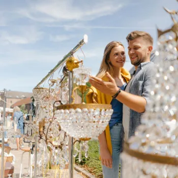 Stel bekijkt kristallen kroonluchters op een zonnige vrijmarkt, met andere bezoekers en kramen op de achtergrond.