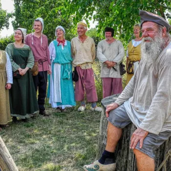 Groep mensen in middeleeuwse kleding tijdens een historisch evenement, met op de voorgrond een man met grijze baard zittend op een boomstam, en op de achtergrond lachende deelnemers in kleurrijke kostuums.