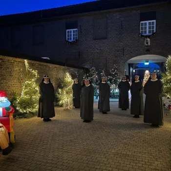 Zusters lopen langs verlichte kerstbomen in de abdijtuin tijdens de Kerststal bij de Zusters in Weert, omgeven door warme lichtjes en kerstdecoraties.