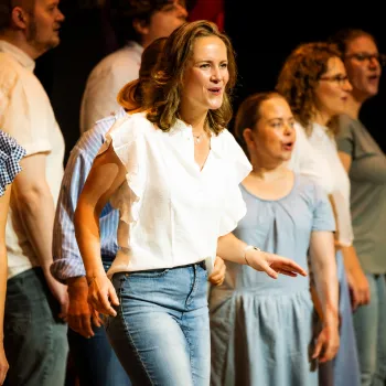Groep zangers treedt samen op tijdens de Limburgse Koordag, met een zangeres in een witte blouse centraal op het podium.