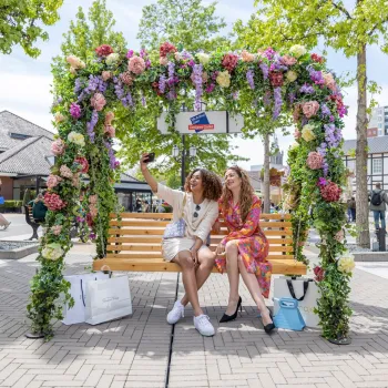 Zwei Frauen sitzen auf einer Holzbank unter einem Blumenbogen und machen ein Selfie während des Mid Season Sale im Designer Outlet Roermond, mit Einkaufstaschen zu ihren Füßen auf einem Platz.