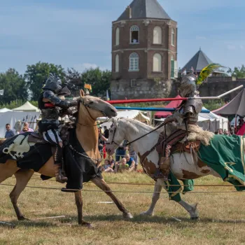 Twee ridders te paard in gevecht tijdens een riddertoernooi bij Kasteel Montfort, omringd door tenten en toeschouwers.
