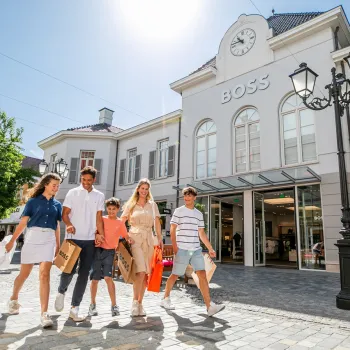 Gezin met shoppingtassen loopt voor de BOSS-winkel tijdens de Summer Sale bij Designer Outlet Roermond op een zonnige dag.