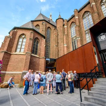 Groep mensen tijdens de VVV Stadswandeling Roermond bij de ingang van de Sint-Christoffelkathedraal in Roermond, met de bakstenen gevel, hoge kerkramen en moderne entree duidelijk in beeld.