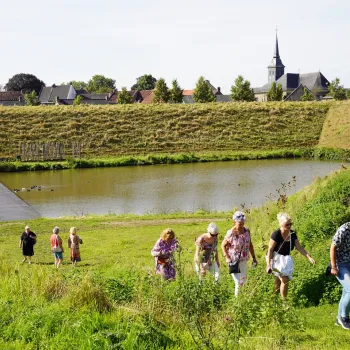 Wandelaars lopen tijdens de VVV wandeling Stevensweert, vesting aan de Maas over een groene dijk langs het water bij Stevensweert, met op de achtergrond een dorp en kerktoren.