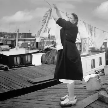 Een voortvarende vrouw hangt de was op tussen woonboten, terwijl ze op het dek staat met klompen aan – een alledaags tafereel uit het leven aan boord.