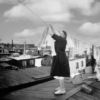 Een voortvarende vrouw hangt de was op tussen woonboten, terwijl ze op het dek staat met klompen aan – een alledaags tafereel uit het leven aan boord.
