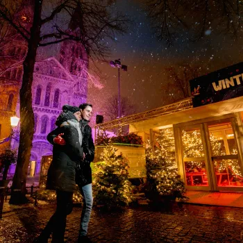A couple walks through the snow past a brightly lit winter pavilion on the Munsterplein in Roermond.
